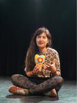 A woman is sitting cross-legged on the floor holding up a heart made from variously coloured carton. She is mid speech, explaining something about the heart.