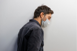 A man with a surgical mask on leaning against a white wall in the background, eyes closed. We see him in profile.