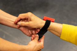 A photo of one person's hands tying a black wristband with a small red box (the movement sensor) to another persons wrist.
